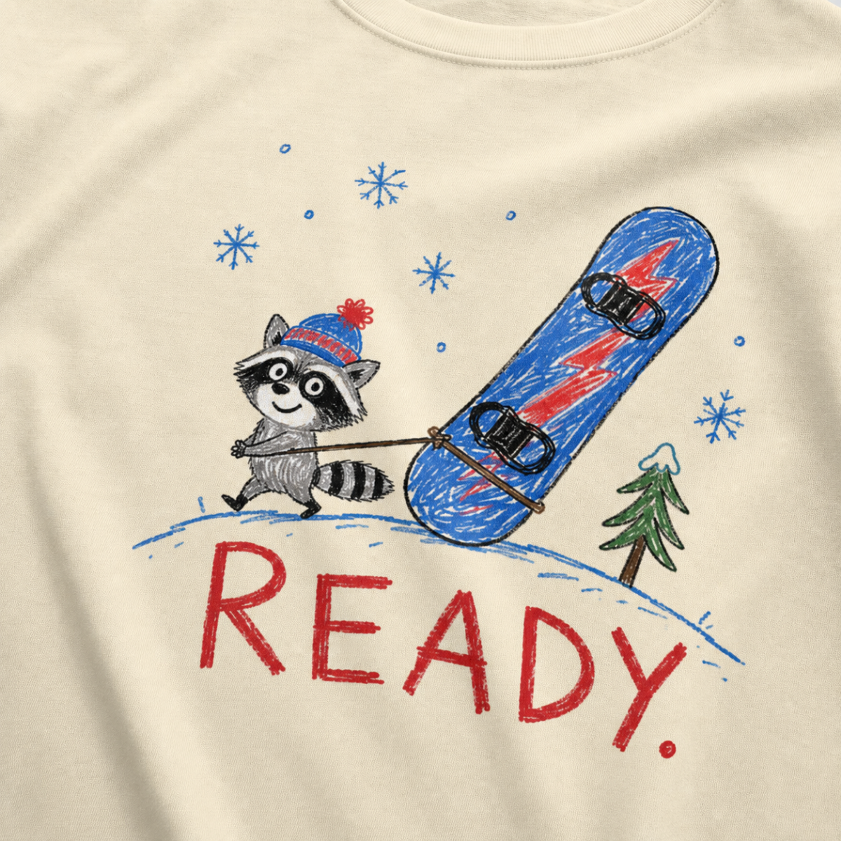 Close-up of an ivory white t-shirt with a playful hand-drawn raccoon snowboarding graphic, snowflakes, a pine tree, and large red “READY.” text.