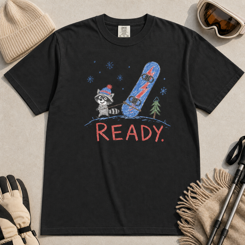 Flat lay of a black t-shirt featuring a childlike crayon-style design with a small raccoon pulling a blue snowboard and bold “READY.” text.