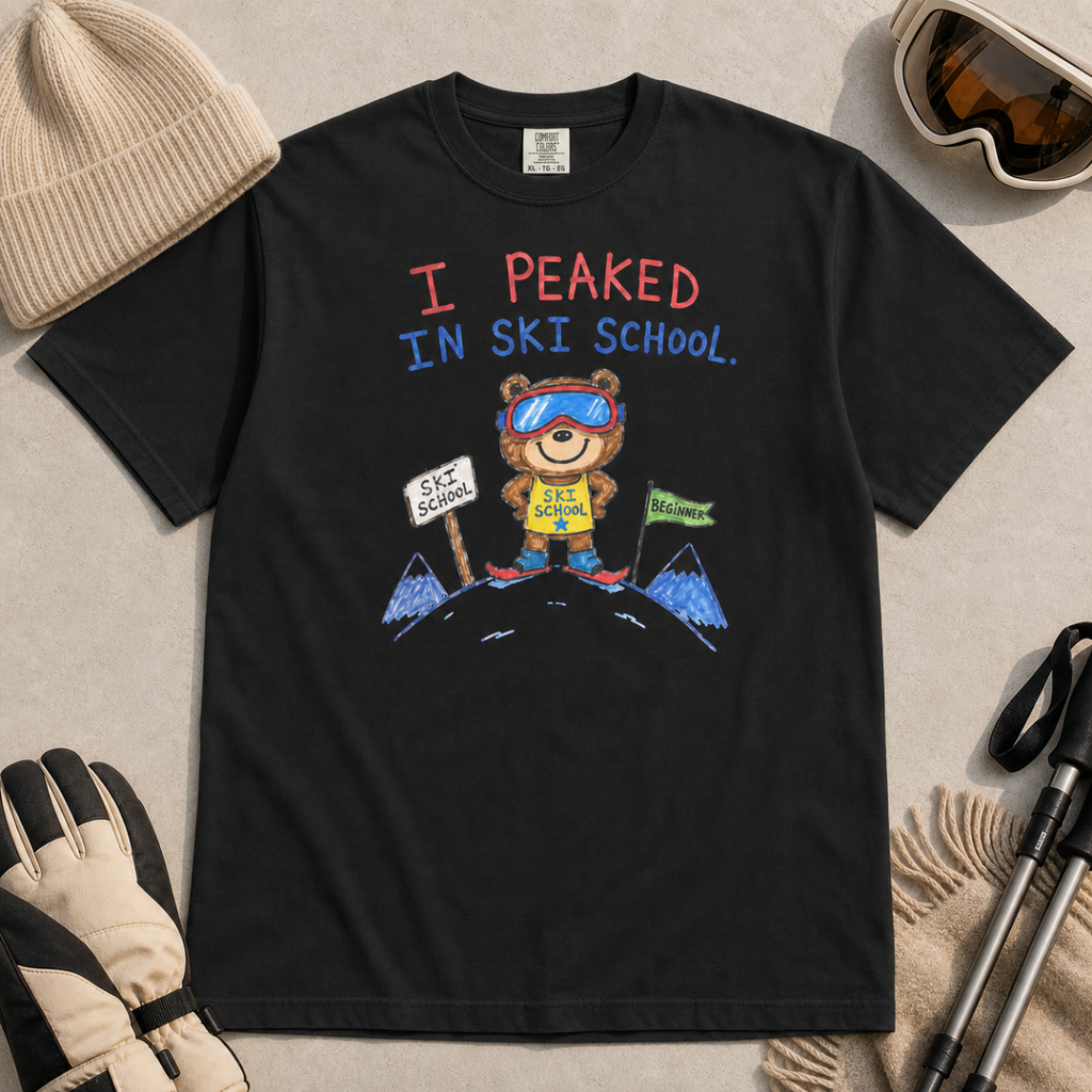 Flat lay of a black t-shirt featuring a childlike crayon-style ski design with a proud bear in ski school gear and “I Peaked in Ski School.” text.