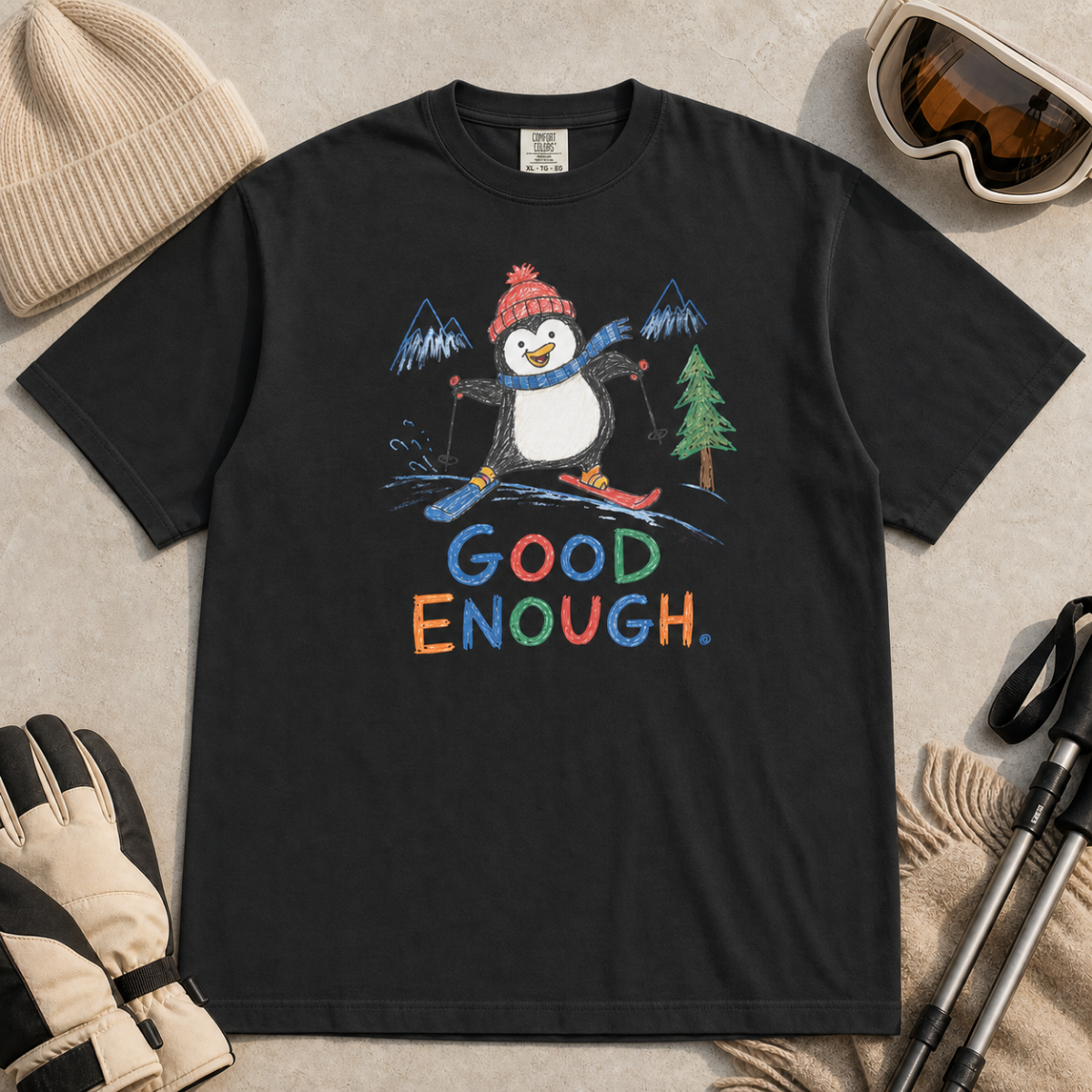 Flat lay of a black t-shirt featuring a childlike crayon-style ski design with a happy penguin skiing downhill, mountains, a pine tree, and colorful “Good Enough.” text.