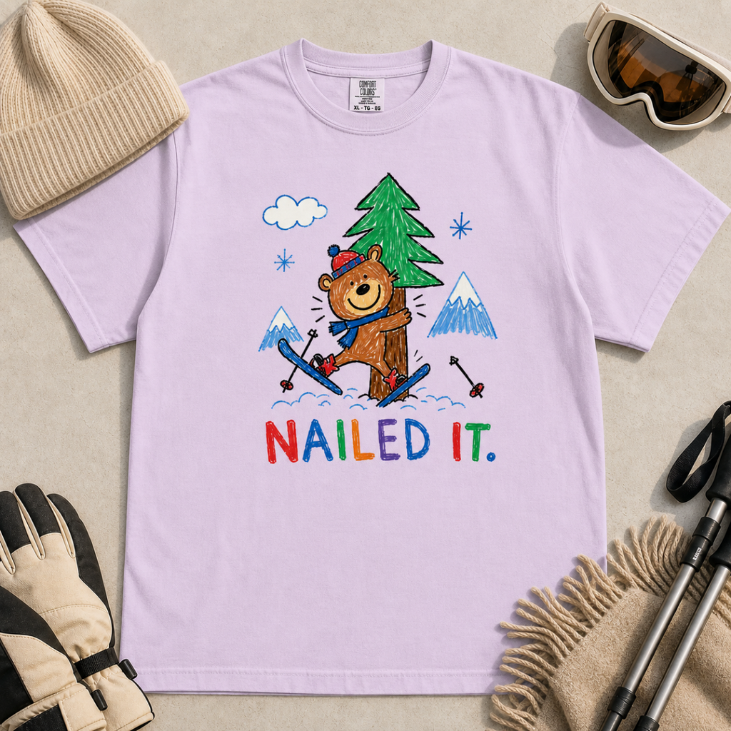 Flat lay of an orchid t-shirt featuring a playful hand-drawn bear ski crash graphic with mountains, snow, a pine tree, and “Nailed It.” text.