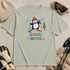 Flat lay of a bay green t-shirt featuring a childlike crayon-style ski design with a happy penguin skiing downhill, mountains, a pine tree, and colorful “Good Enough.” text.