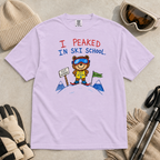 Flat lay of an orchid t-shirt featuring a childlike crayon-style ski design with a proud bear in ski school gear and “I Peaked in Ski School.” text.