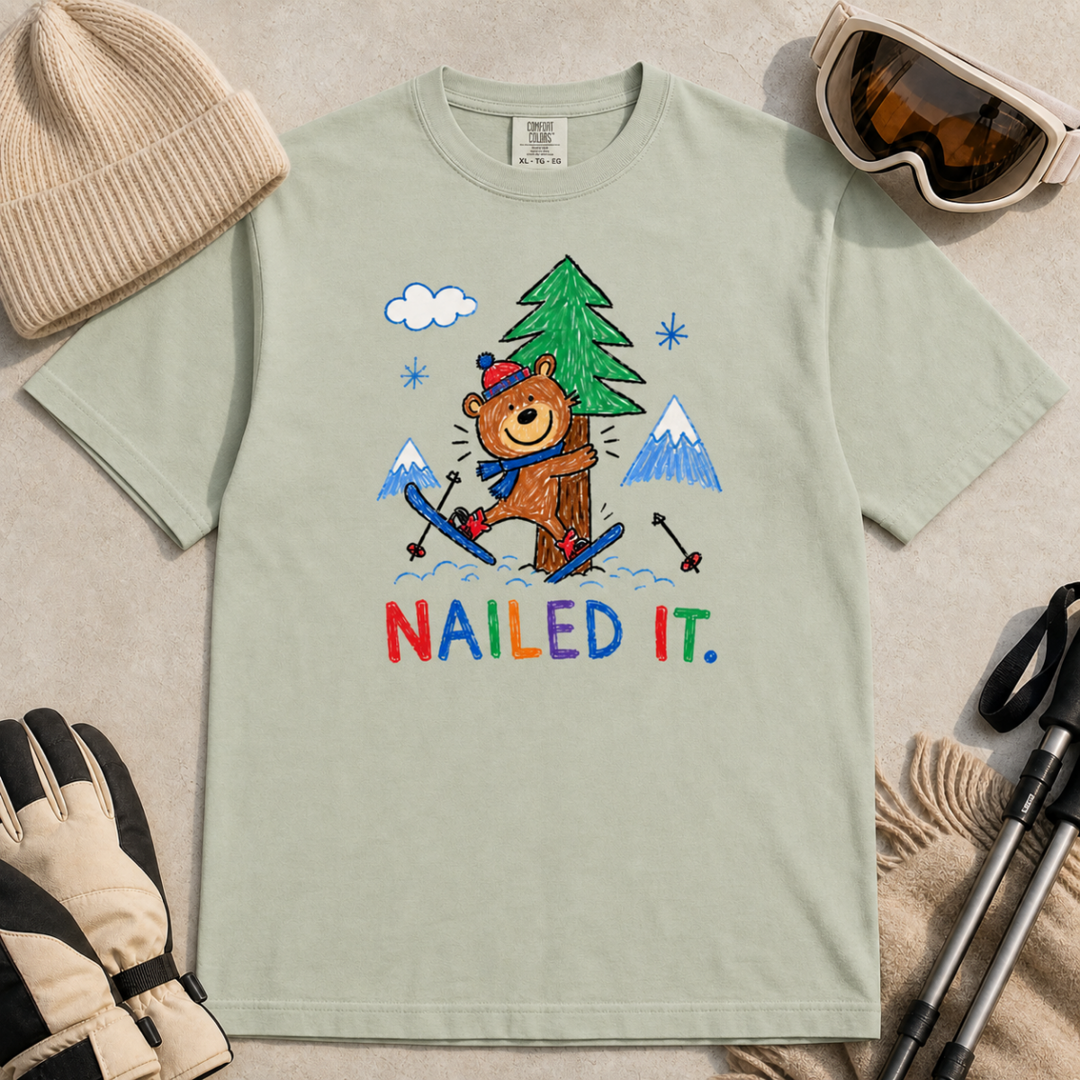 Flat lay of a bay green t-shirt featuring a playful hand-drawn bear ski crash graphic with mountains, snow, a pine tree, and “Nailed It.” text.