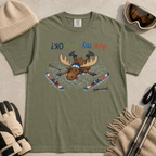 Flat lay of a moss green t-shirt featuring a humorous upside-down “Are My Skis OK?” design with a moose face-planted in the snow among scattered skis and poles.