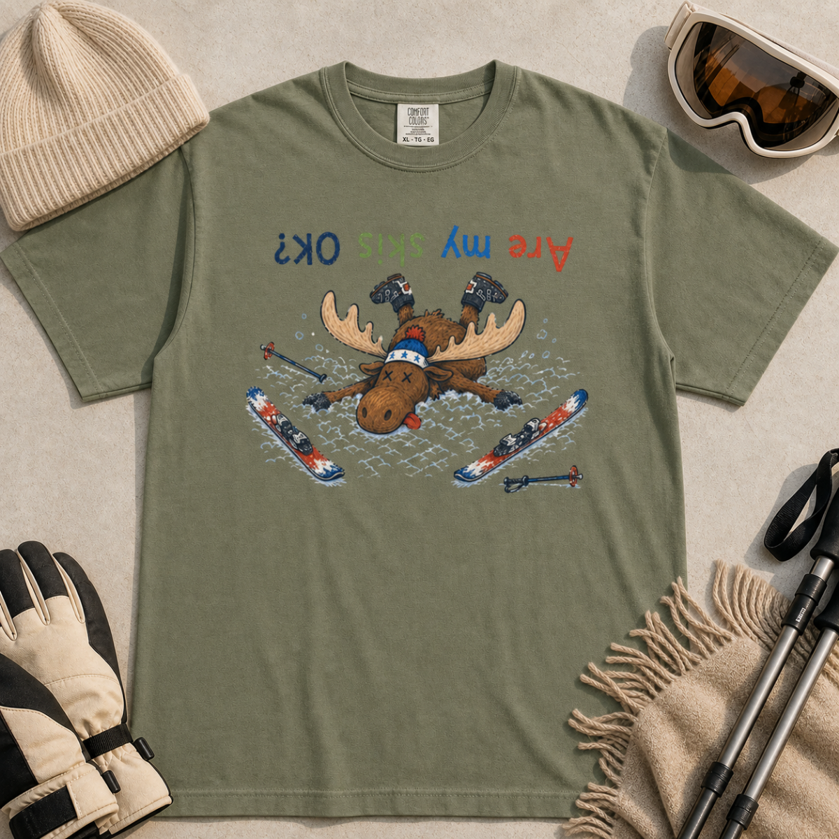Flat lay of a moss green t-shirt featuring a humorous upside-down “Are My Skis OK?” design with a moose face-planted in the snow among scattered skis and poles.