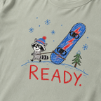 Close-up of a green bay t-shirt with a playful hand-drawn raccoon snowboarding graphic, snowflakes, a pine tree, and large red “READY.” text.