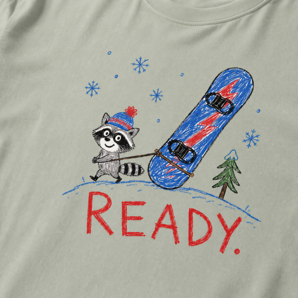Close-up of a green bay t-shirt with a playful hand-drawn raccoon snowboarding graphic, snowflakes, a pine tree, and large red “READY.” text.