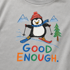 Close-up of a granite gray t-shirt with a playful hand-drawn penguin ski graphic, snowy mountain details, and large colorful “Good Enough.” text.