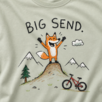 Close-up of a bay green t-shirt with a childlike crayon-style mountain biking design showing a joyful fox standing on a dirt jump with “Big Send.” text.