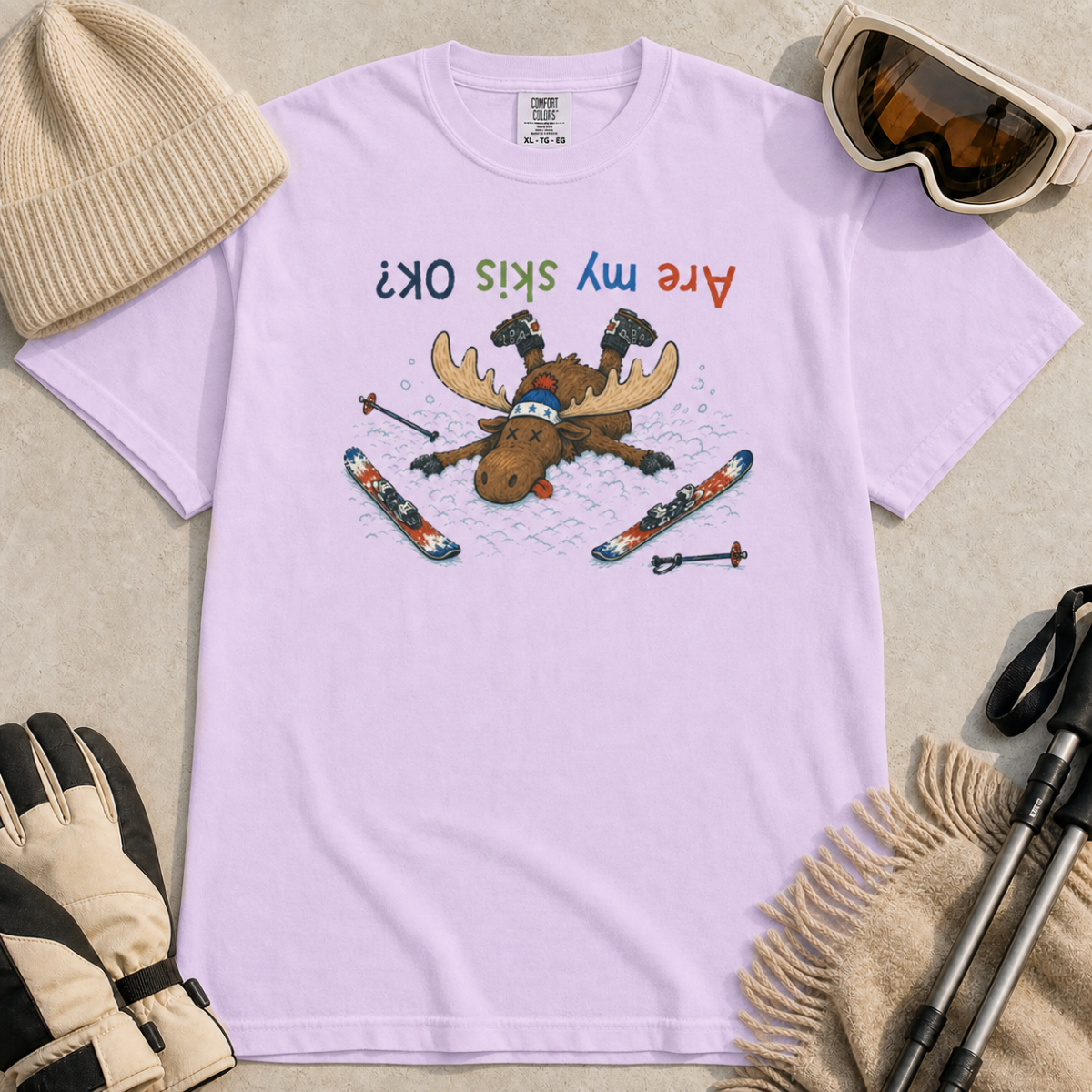 Flat lay of an orchid t-shirt featuring a humorous upside-down “Are My Skis OK?” design with a moose face-planted in the snow among scattered skis and poles.