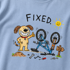 Close-up of a washed denim blue t-shirt with a playful hand-drawn dog giving a thumbs up beside a repaired upside-down bike, scattered tools, and “FIXED.” text.