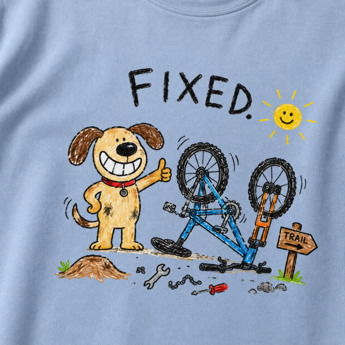 Close-up of a washed denim blue t-shirt with a playful hand-drawn dog giving a thumbs up beside a repaired upside-down bike, scattered tools, and “FIXED.” text.
