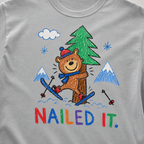 Close-up of a granite gray t-shirt with a childlike crayon-style ski design showing a smiling bear hugging a tree, with colorful “Nailed It.” text.