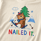 Close-up of an ivory white t-shirt with a childlike crayon-style ski design showing a smiling bear hugging a tree, with colorful “Nailed It.” text.