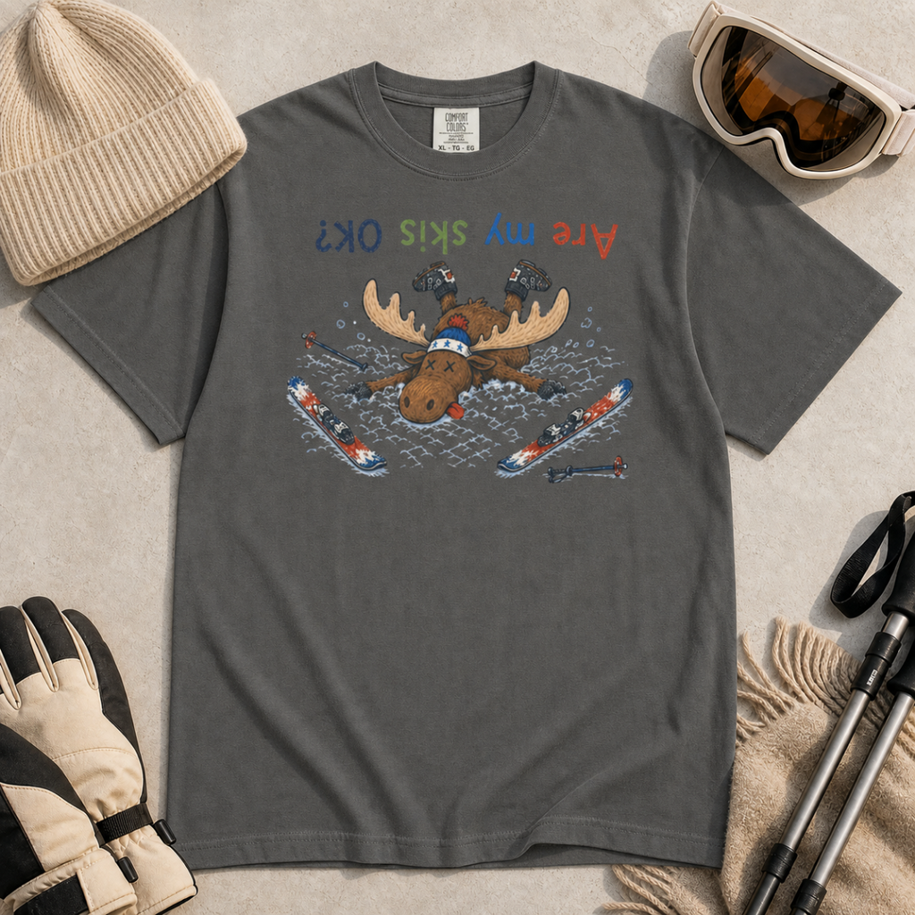 Flat lay of a pepper gray t-shirt featuring a humorous upside-down “Are My Skis OK?” design with a moose face-planted in the snow among scattered skis and poles.