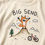 Close-up of an ivory white t-shirt with a childlike crayon-style mountain biking design showing a joyful fox standing on a dirt jump with “Big Send.” text.