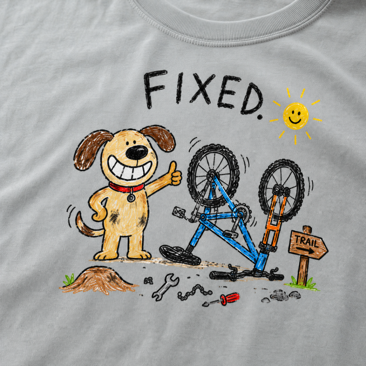 Close-up of a granite gray t-shirt with a playful hand-drawn dog giving a thumbs up beside a repaired upside-down bike, scattered tools, and “FIXED.” text.