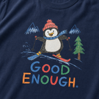 Close-up of a navy t-shirt with a playful hand-drawn penguin ski graphic, snowy mountain details, and large colorful “Good Enough.” text.