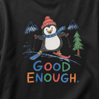 Close-up of a black t-shirt with a playful hand-drawn penguin ski graphic, snowy mountain details, and large colorful “Good Enough.” text.