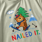 Close-up of a bay green t-shirt with a childlike crayon-style ski design showing a smiling bear hugging a tree, with colorful “Nailed It.” text.