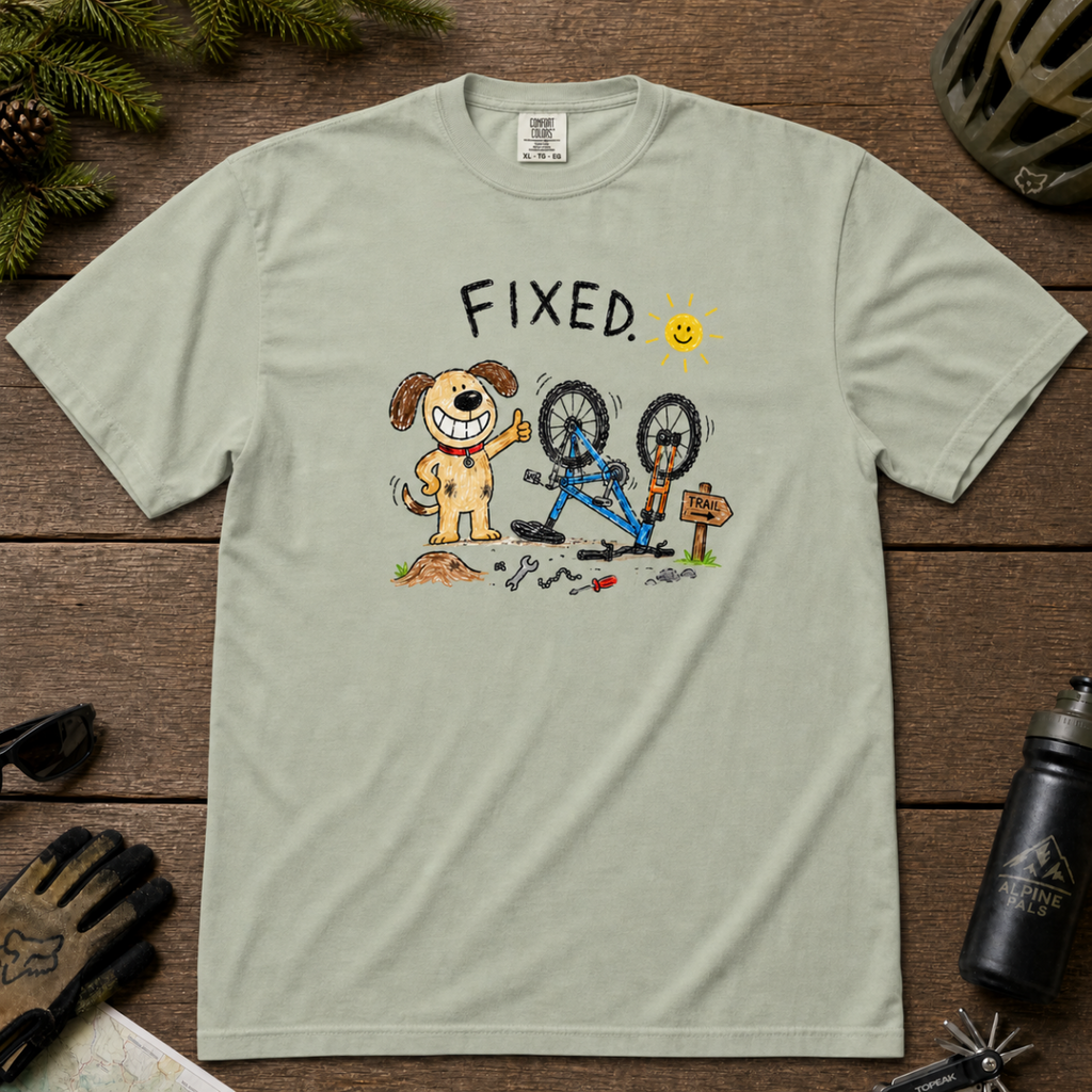 Flat lay of a bay green t-shirt featuring a childlike crayon-style bike repair design with a smiling dog, upside-down mountain bike, tools, and “FIXED.” text.