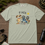 Flat lay of a bay green t-shirt featuring a childlike crayon-style bike repair design with a smiling dog, upside-down mountain bike, tools, and “FIXED.” text.