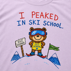 Close-up of an orchid t-shirt with a playful hand-drawn bear at ski school, beginner flag, mountain details, and “I Peaked in Ski School.” text.