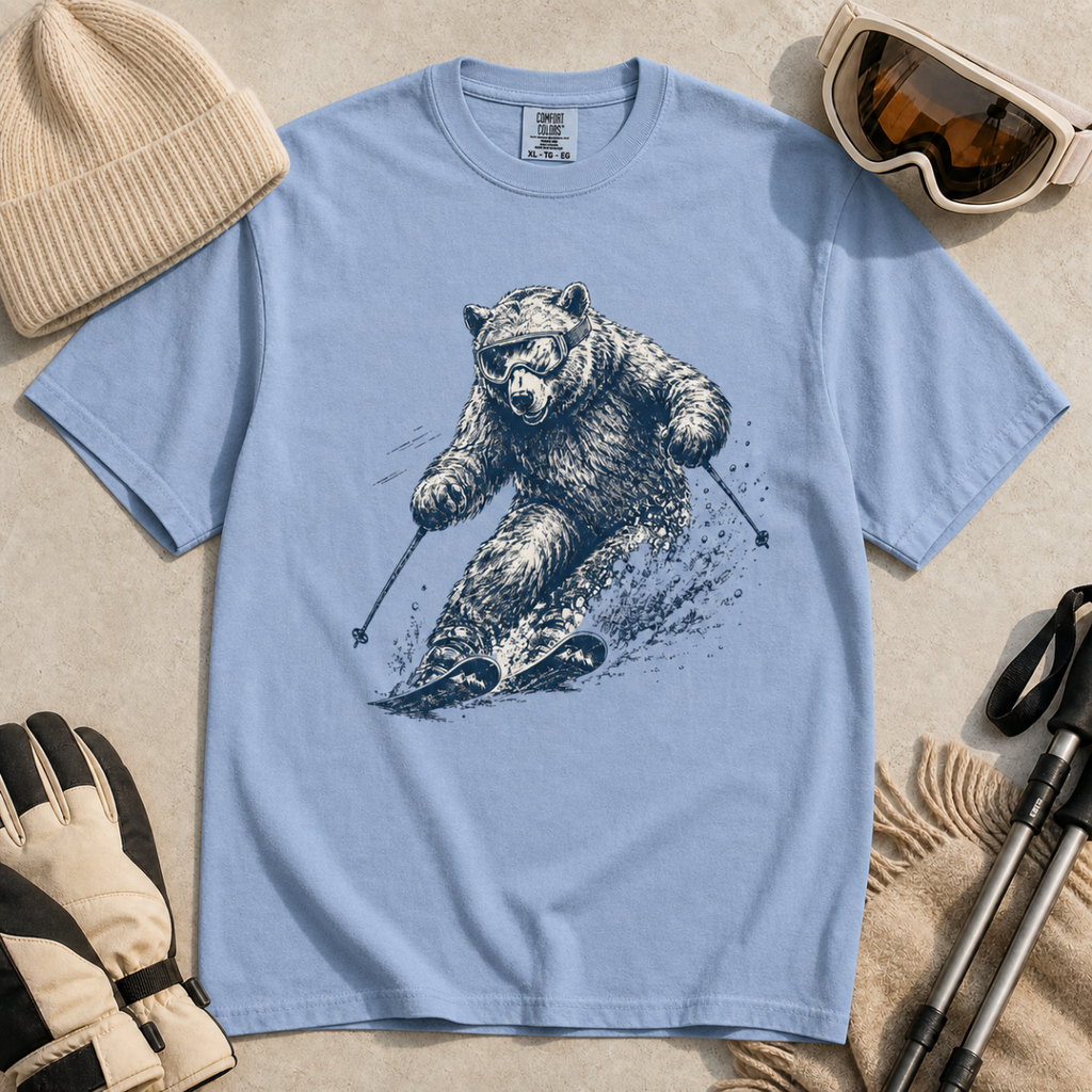 Flat lay of a washed denim blue t-shirt featuring a detailed blue line-art bear skiing through powder, styled with winter accessories.