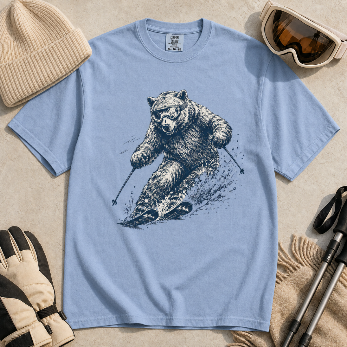 Flat lay of a washed denim blue t-shirt featuring a detailed blue line-art bear skiing through powder, styled with winter accessories.