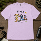 Flat lay of an orchid t-shirt featuring a childlike crayon-style bike repair design with a smiling dog, upside-down mountain bike, tools, and “FIXED.” text.