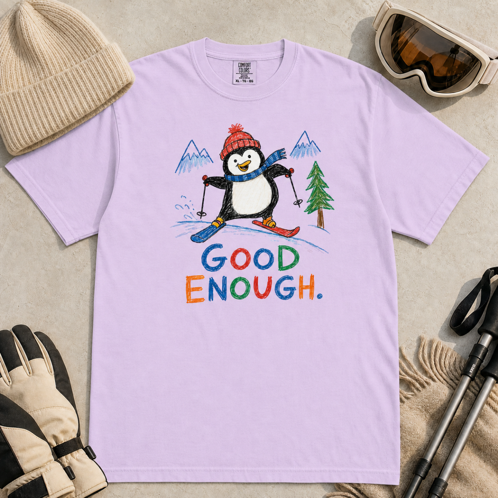 Flat lay of an orchid t-shirt featuring a childlike crayon-style ski design with a happy penguin skiing downhill, mountains, a pine tree, and colorful “Good Enough.” text.