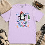 Flat lay of an orchid t-shirt featuring a childlike crayon-style ski design with a happy penguin skiing downhill, mountains, a pine tree, and colorful “Good Enough.” text.