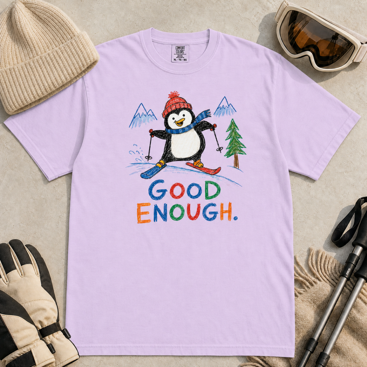 Flat lay of an orchid t-shirt featuring a childlike crayon-style ski design with a happy penguin skiing downhill, mountains, a pine tree, and colorful “Good Enough.” text.