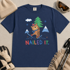 Flat lay of a black t-shirt featuring a playful hand-drawn bear ski crash graphic with mountains, snow, a pine tree, and “Nailed It.” text.