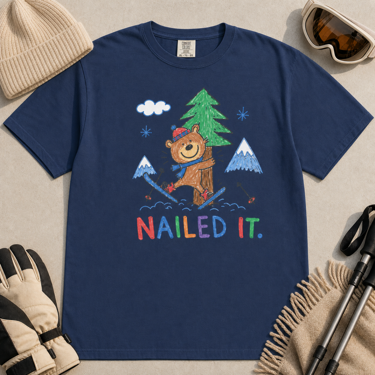 Flat lay of a black t-shirt featuring a playful hand-drawn bear ski crash graphic with mountains, snow, a pine tree, and “Nailed It.” text.