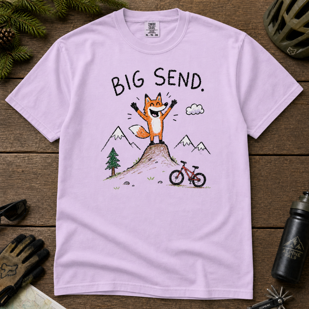 Flat lay of an orchid t-shirt featuring a playful hand-drawn fox mountain bike graphic with mountains, a small bike, and “Big Send.” text.