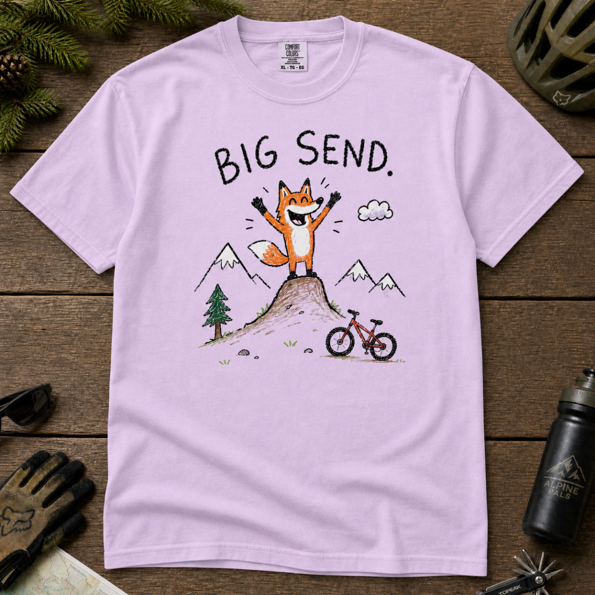 Flat lay of an orchid t-shirt featuring a playful hand-drawn fox mountain bike graphic with mountains, a small bike, and “Big Send.” text.