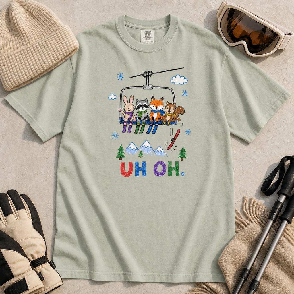 Flat lay of a bay green t-shirt featuring a playful hand-drawn chairlift scene with bunny, raccoon, fox, and squirrel skiers, snowy mountains, and “UH OH.” text.