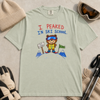 Flat lay of a bay green t-shirt featuring a childlike crayon-style ski design with a proud bear in ski school gear and “I Peaked in Ski School.” text.