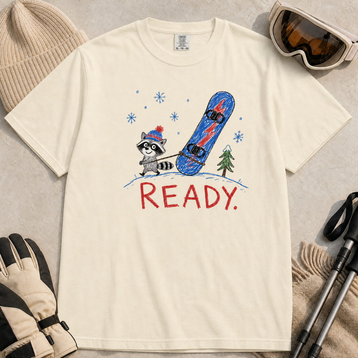 Flat lay of an ivory white t-shirt featuring a childlike crayon-style design with a small raccoon pulling a blue snowboard and bold “READY.” text.