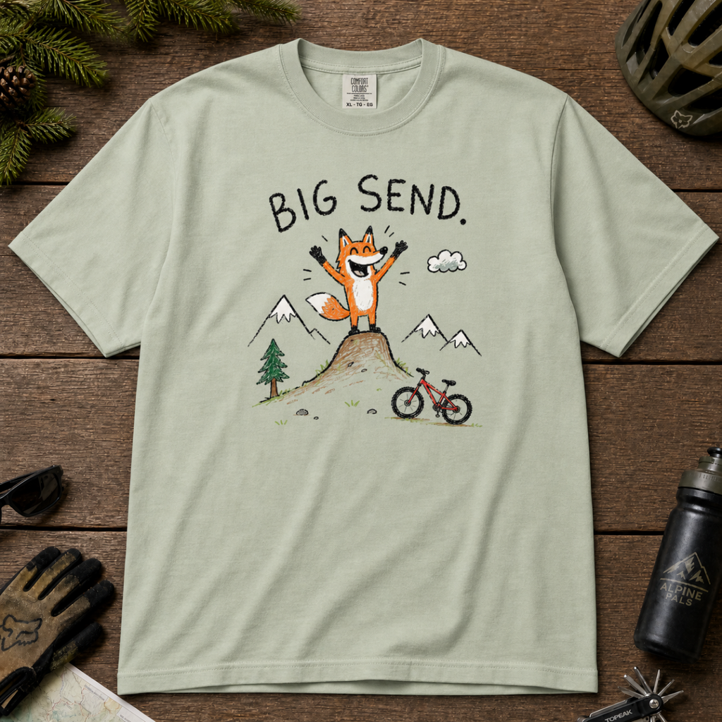 Flat lay of a bay green t-shirt featuring a playful hand-drawn fox mountain bike graphic with mountains, a small bike, and “Big Send.” text.