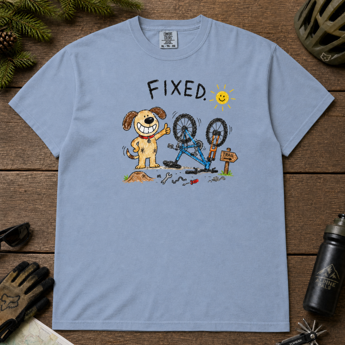 Flat lay of a washed denim blue t-shirt featuring a childlike crayon-style bike repair design with a smiling dog, upside-down mountain bike, tools, and “FIXED.” text.