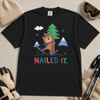 Flat lay of a black t-shirt featuring a playful hand-drawn bear ski crash graphic with mountains, snow, a pine tree, and “Nailed It.” text.