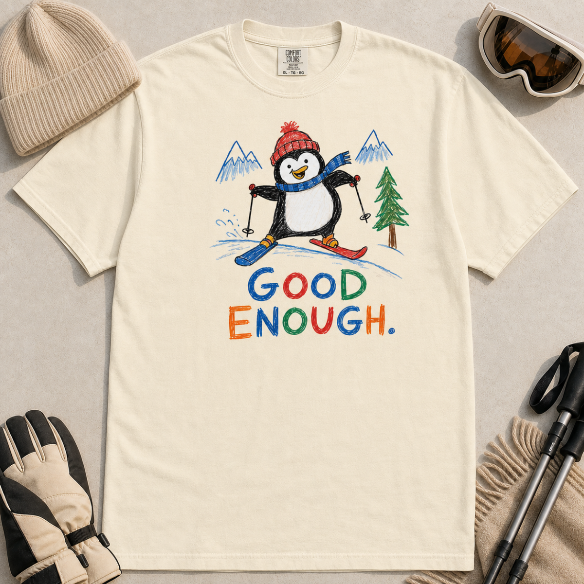 Flat lay of an ivory white t-shirt featuring a childlike crayon-style ski design with a happy penguin skiing downhill, mountains, a pine tree, and colorful “Good Enough.” text.