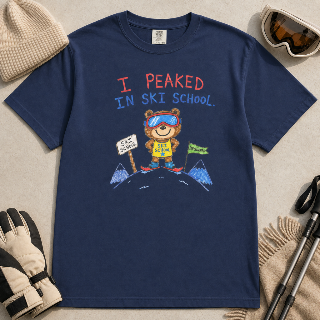 Flat lay of a navy t-shirt featuring a childlike crayon-style ski design with a proud bear in ski school gear and “I Peaked in Ski School.” text.
