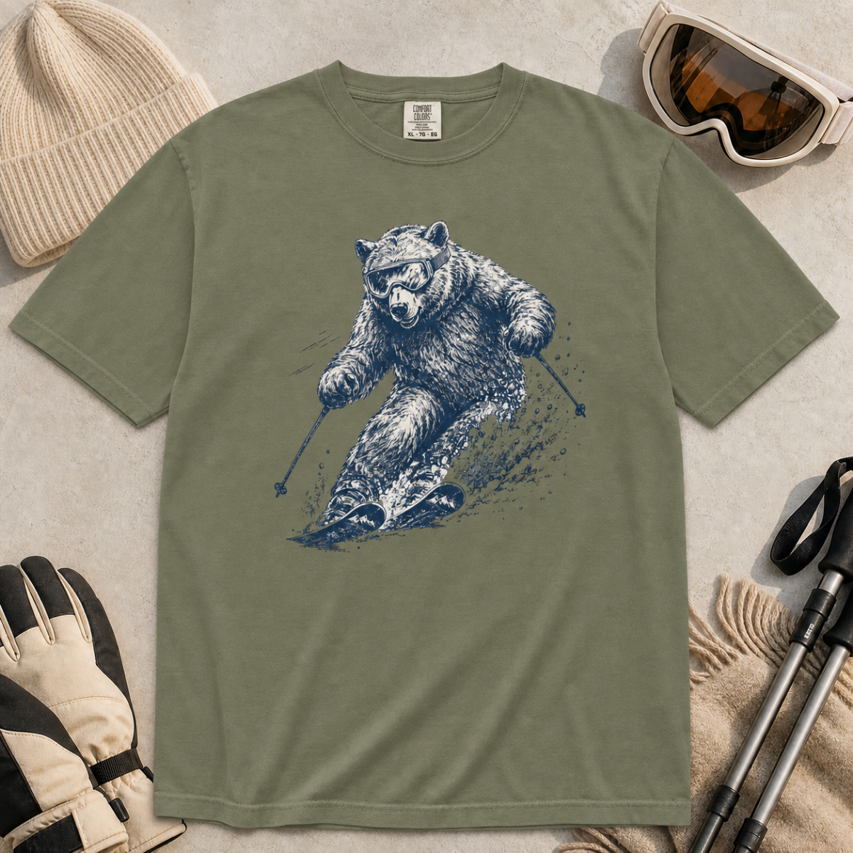 Flat lay of a moss green t-shirt featuring a detailed blue line-art bear skiing through powder, styled with winter accessories.
