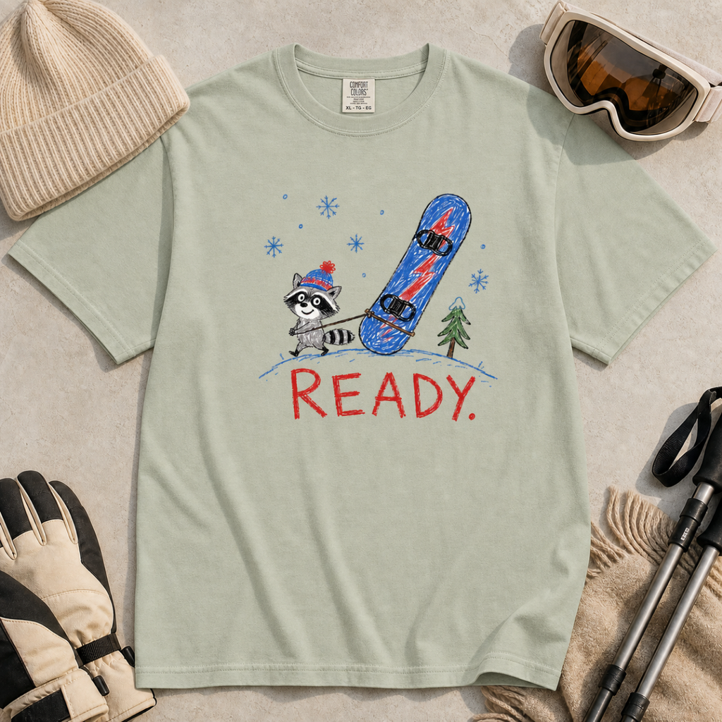Flat lay of a bay green t-shirt featuring a childlike crayon-style design with a small raccoon pulling a blue snowboard and bold “READY.” text.