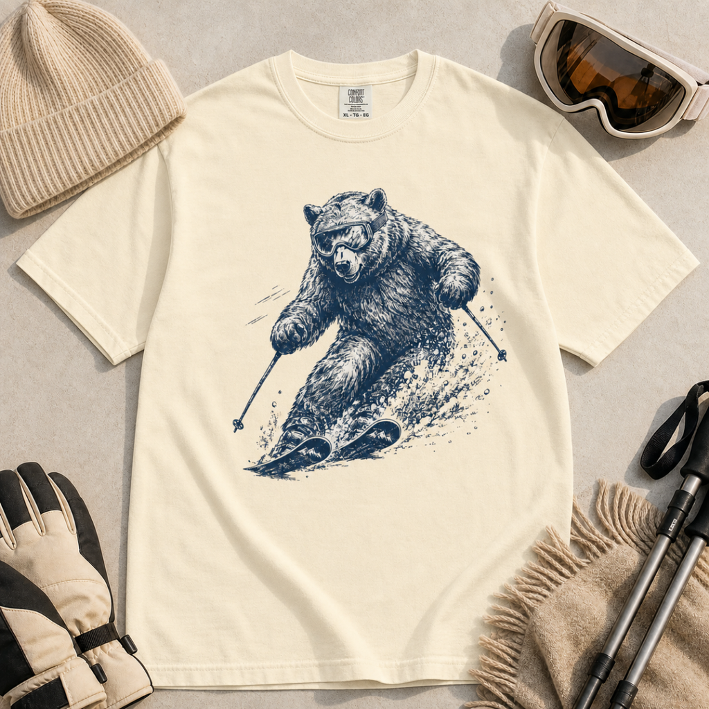 Flat lay of an ivory white t-shirt featuring a detailed blue line-art bear skiing through powder, styled with winter accessories.