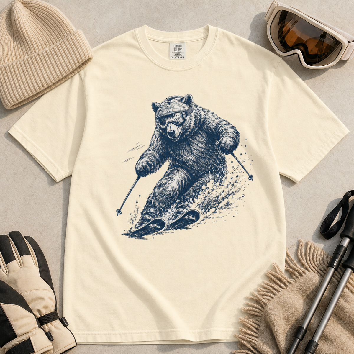 Flat lay of an ivory white t-shirt featuring a detailed blue line-art bear skiing through powder, styled with winter accessories.